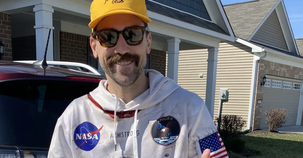 Mark Titus standing outside his house wearing NASA hoodie and yellow cap with residential neighborhood in background